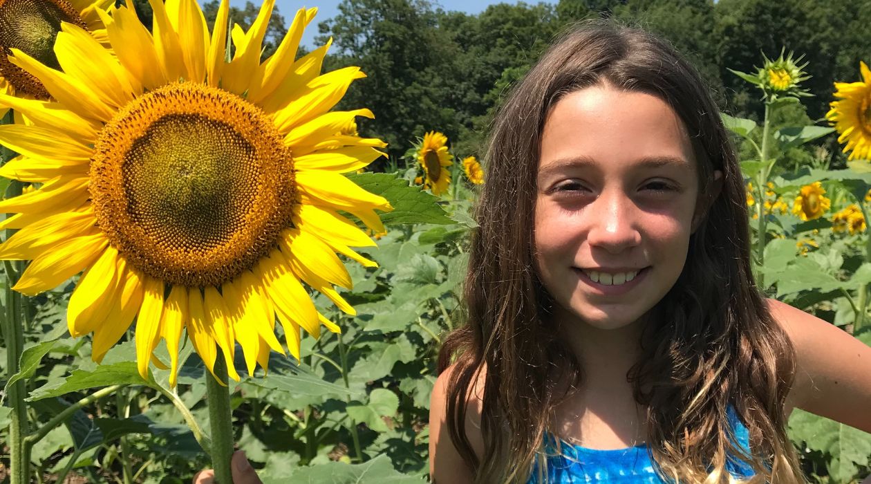 A young girl wearing a bright blue shirt smiles as she holds a big, yellow sunflower at Hudson Valley Cold Pressed Oils in Pleasant Valley.